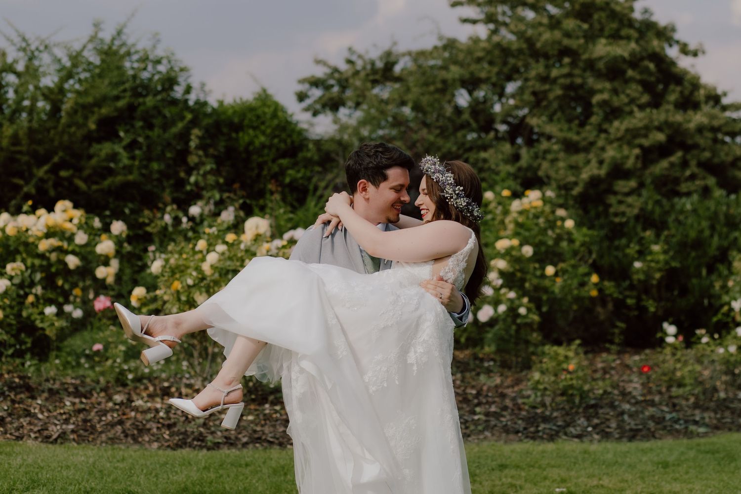 A couple in wedding attire walks hand in hand across a grassy field at sunset with dramatic clouds overhead.