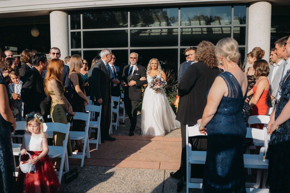 Wedding guests seated in white chairs at an outdoor ceremony with stone columns and blue skies.