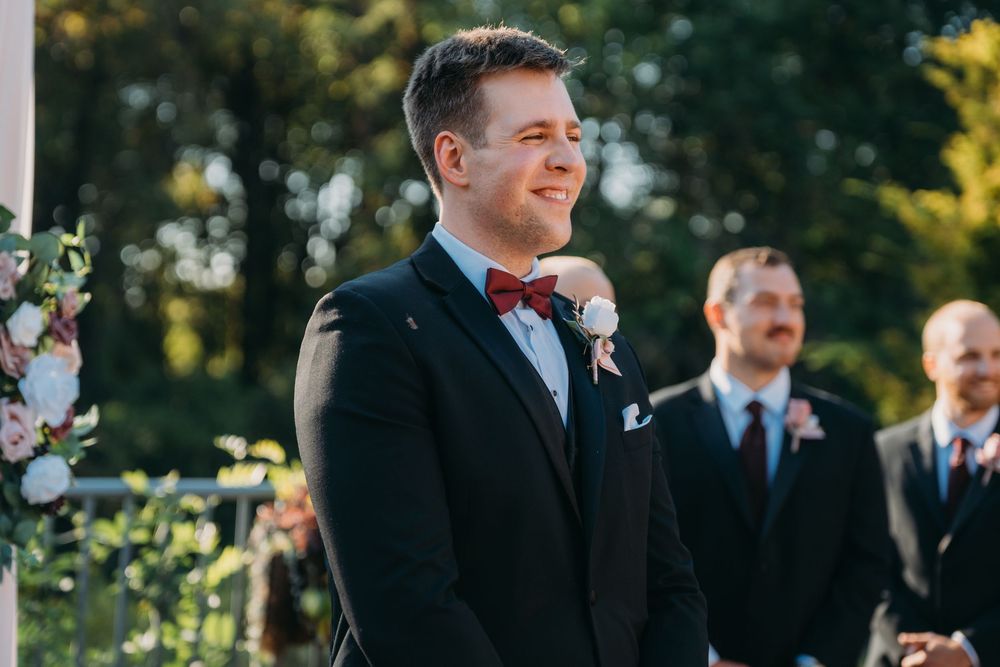 Groom wearing black tuxedo and red bowtie smiles joyfully during outdoor wedding ceremony with groomsmen in background.