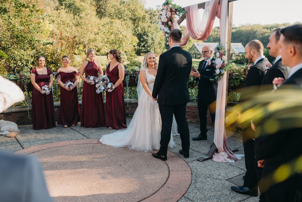 Wedding party members gather outdoors on a paved courtyard during sunset ceremony with burgundy attire.