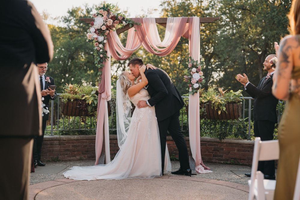 A romantic wedding ceremony takes place under a draped floral arch with pink and white decorations in an outdoor garden setting.