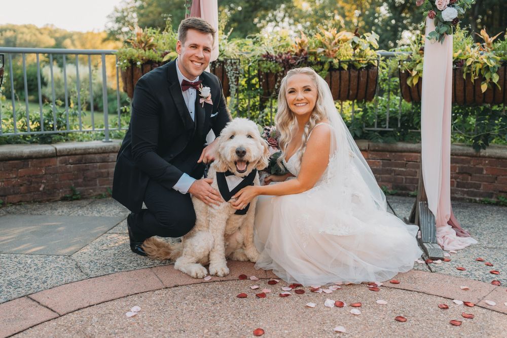 Newlyweds pose with their fluffy white dog in a garden setting with brick pathways.