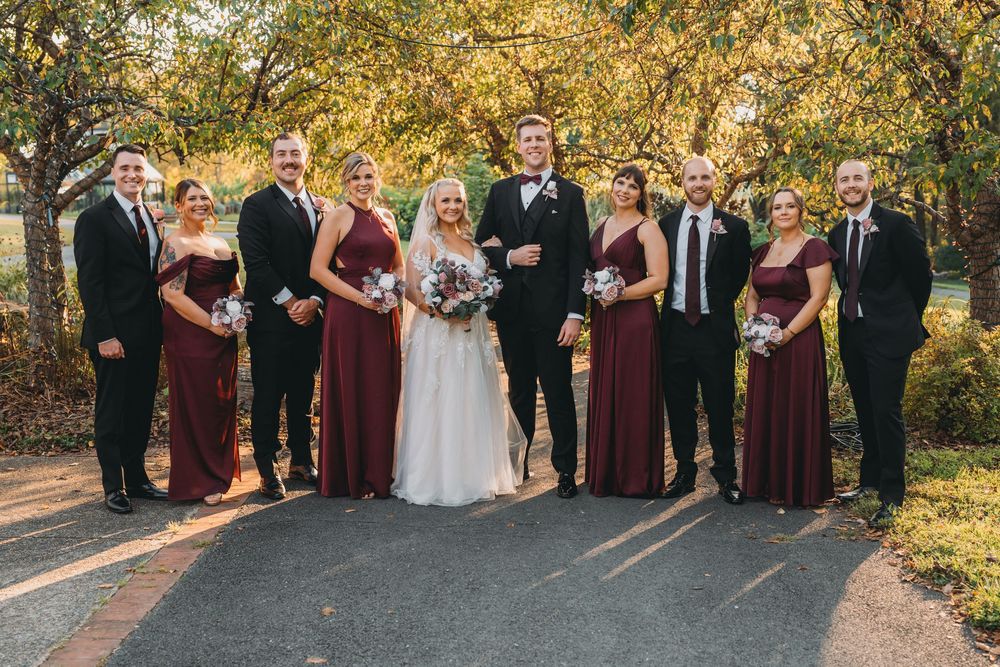 Wedding party wearing burgundy bridesmaid dresses and black suits poses on paved path during autumn season.