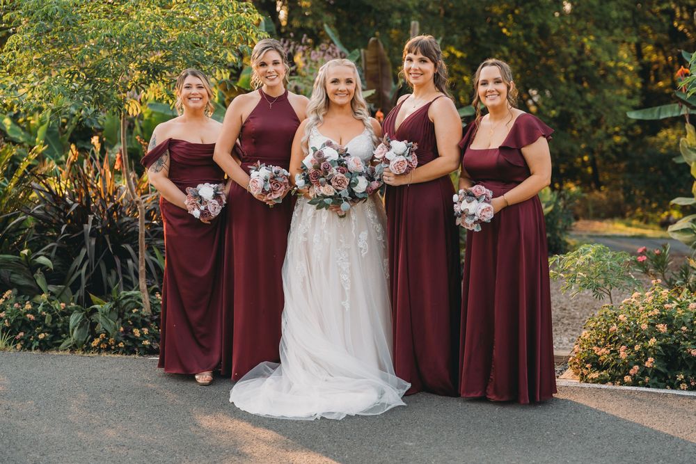 Wedding party wearing burgundy dresses and holding floral bouquets pose together in an autumn garden setting.