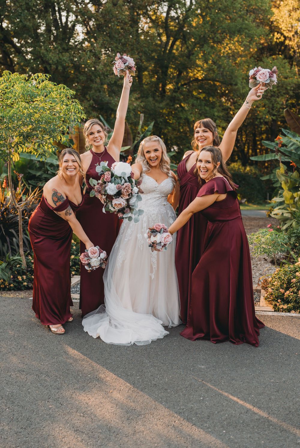 A bridal party in burgundy dresses and white gown poses joyfully on a garden path at sunset at the Atrium at Meadowlark Botanical Garden