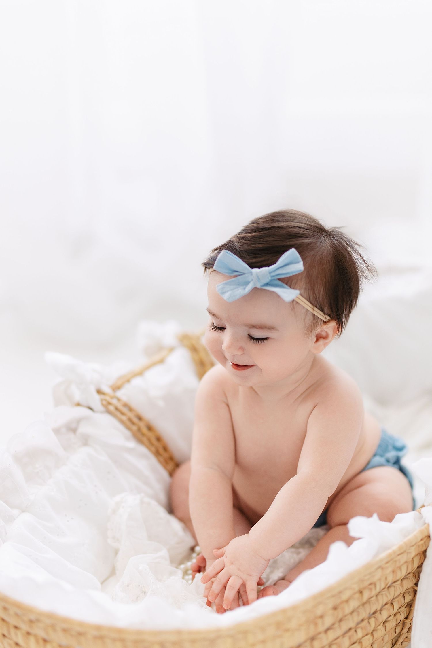 A baby in a blue bow hairband lies peacefully on white bedding in a woven basket.