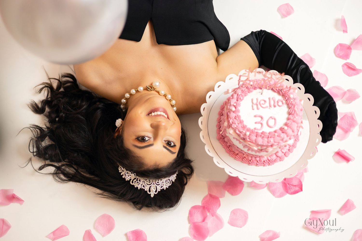 A person lying down surrounded by pink rose petals and decorative cakes in a romantic styled photo shoot.