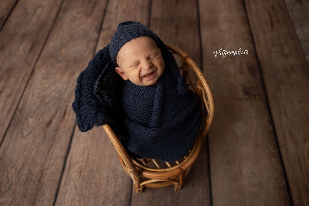 A baby wearing a navy blue knit hat smiles while lying on a rustic wooden floor.