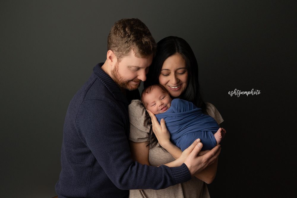 A tender family portrait against a dark background showing parents embracing their newborn baby.