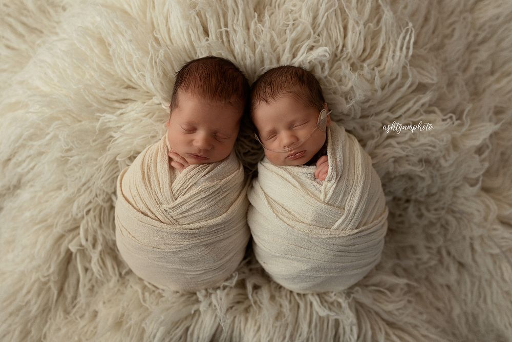 Professional newborn twin photography with babies wrapped in cream fabrics on soft furry backdrop.