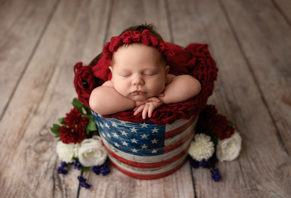Sleeping newborn nestled in patriotic American flag container surrounded by red and white flowers on wooden floor.