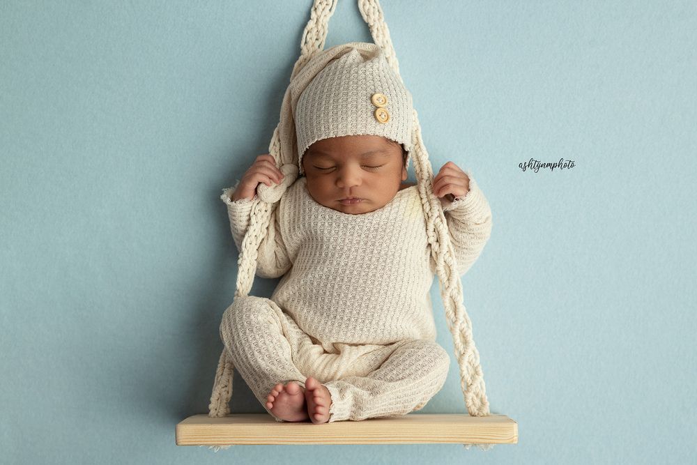 Sleeping newborn in cozy white knit outfit rests peacefully on small wooden swing against blue background.