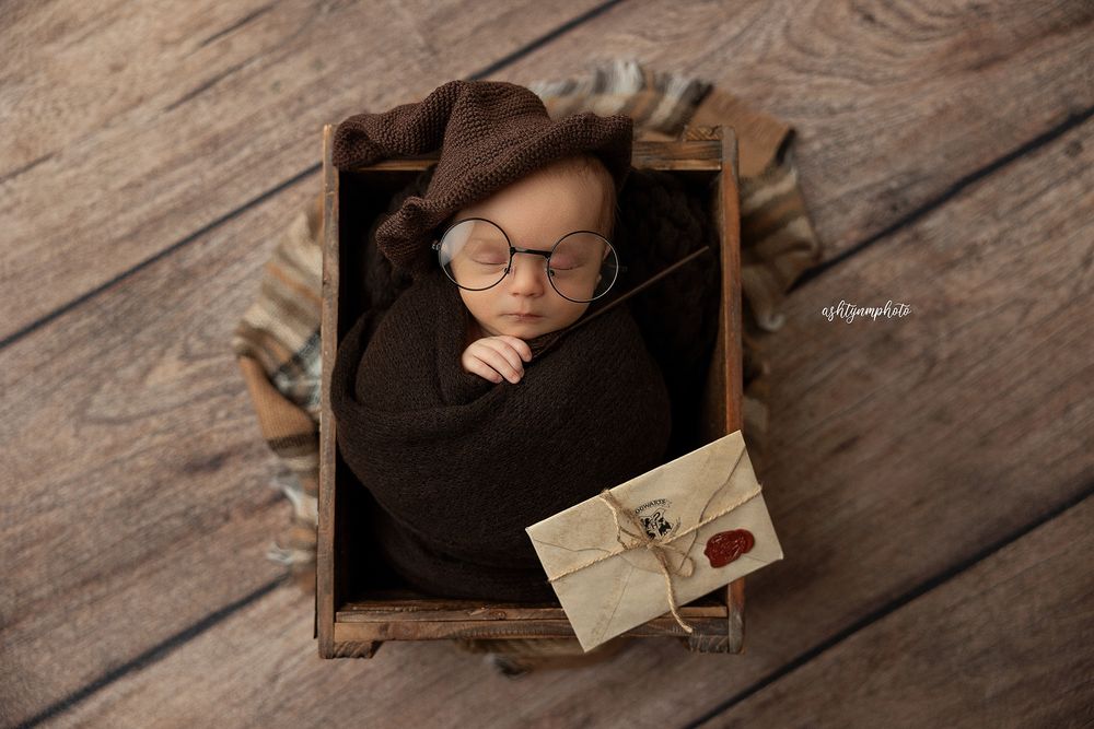 A sleeping newborn in round glasses nestled in a wooden box with a vintage letter prop.