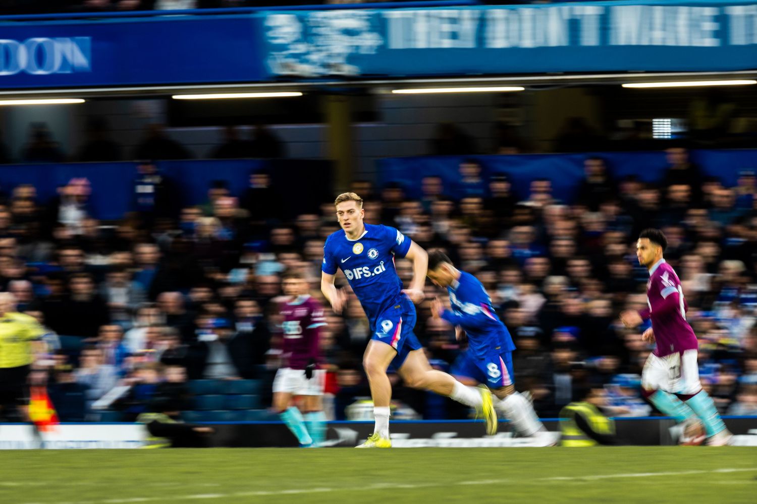 A rugby player in blue jersey carries the ball while running during a match at a stadium.