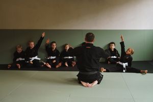 Students in black martial arts uniforms sitting in a row while instructor demonstrates technique.