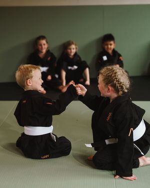 Young martial arts students in black uniforms practicing techniques while sitting on training mats.