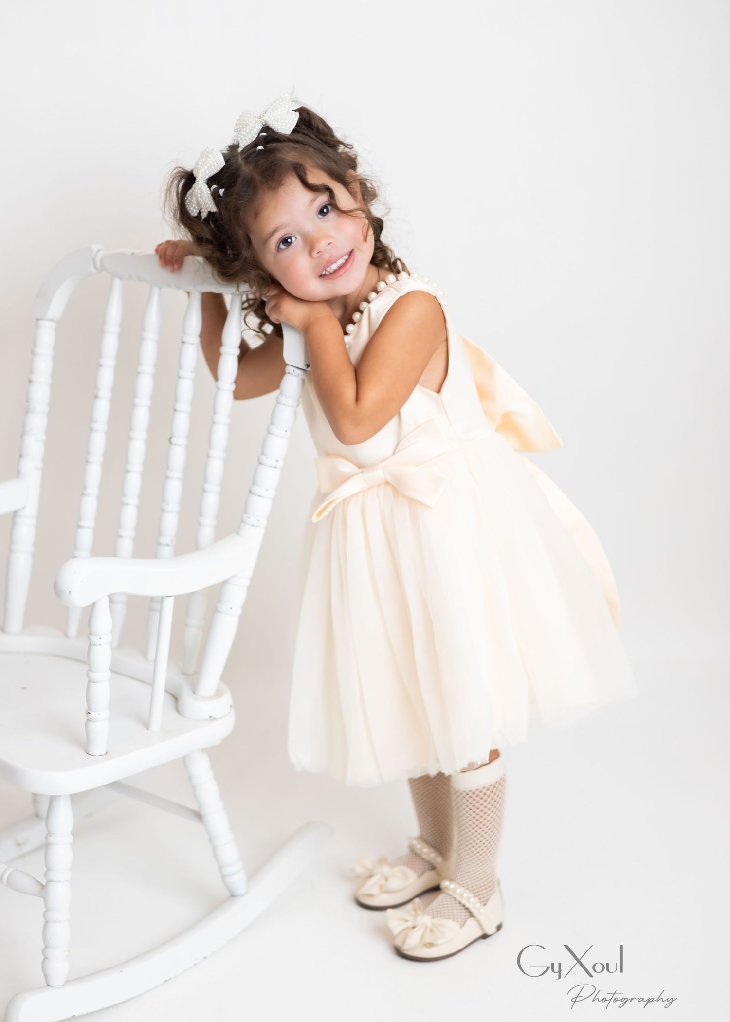 A child in a cream-colored dress poses playfully near a white rocking chair in a bright studio setting.