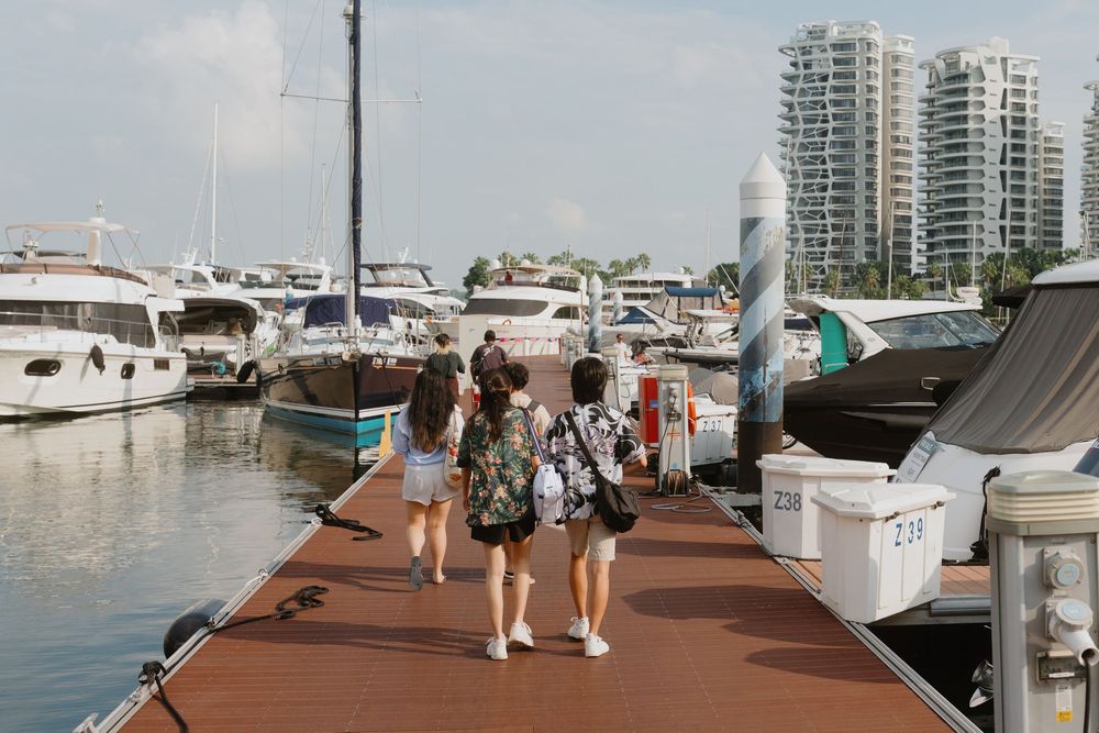People walking along a marina dock with luxury yachts and high-rise buildings in the background.