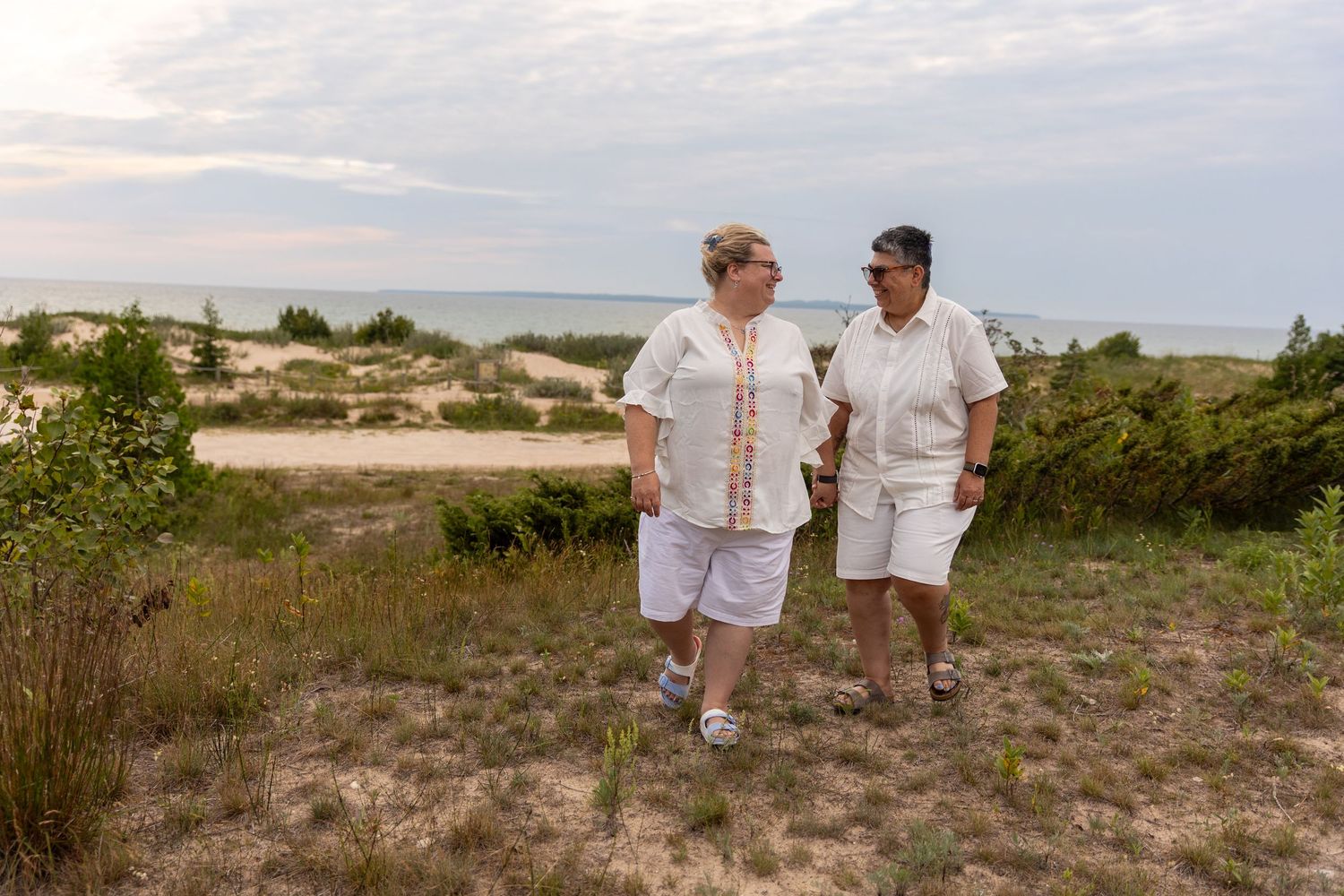 Two women walking together on a sandy coastal trail with ocean views and green dunes in the background.