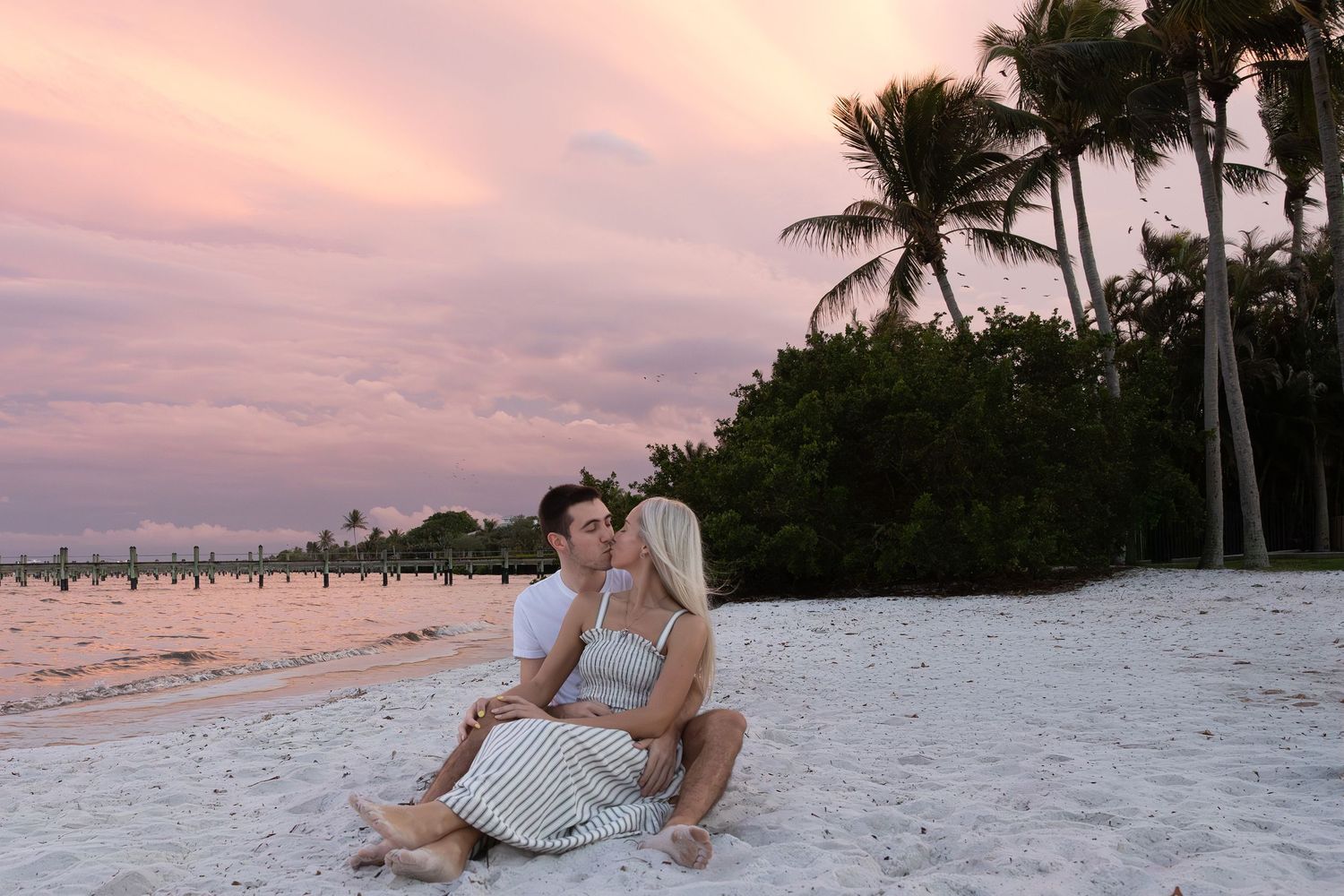 Couple sitting together on a white sandy beach at sunset with pink sky and palm trees.