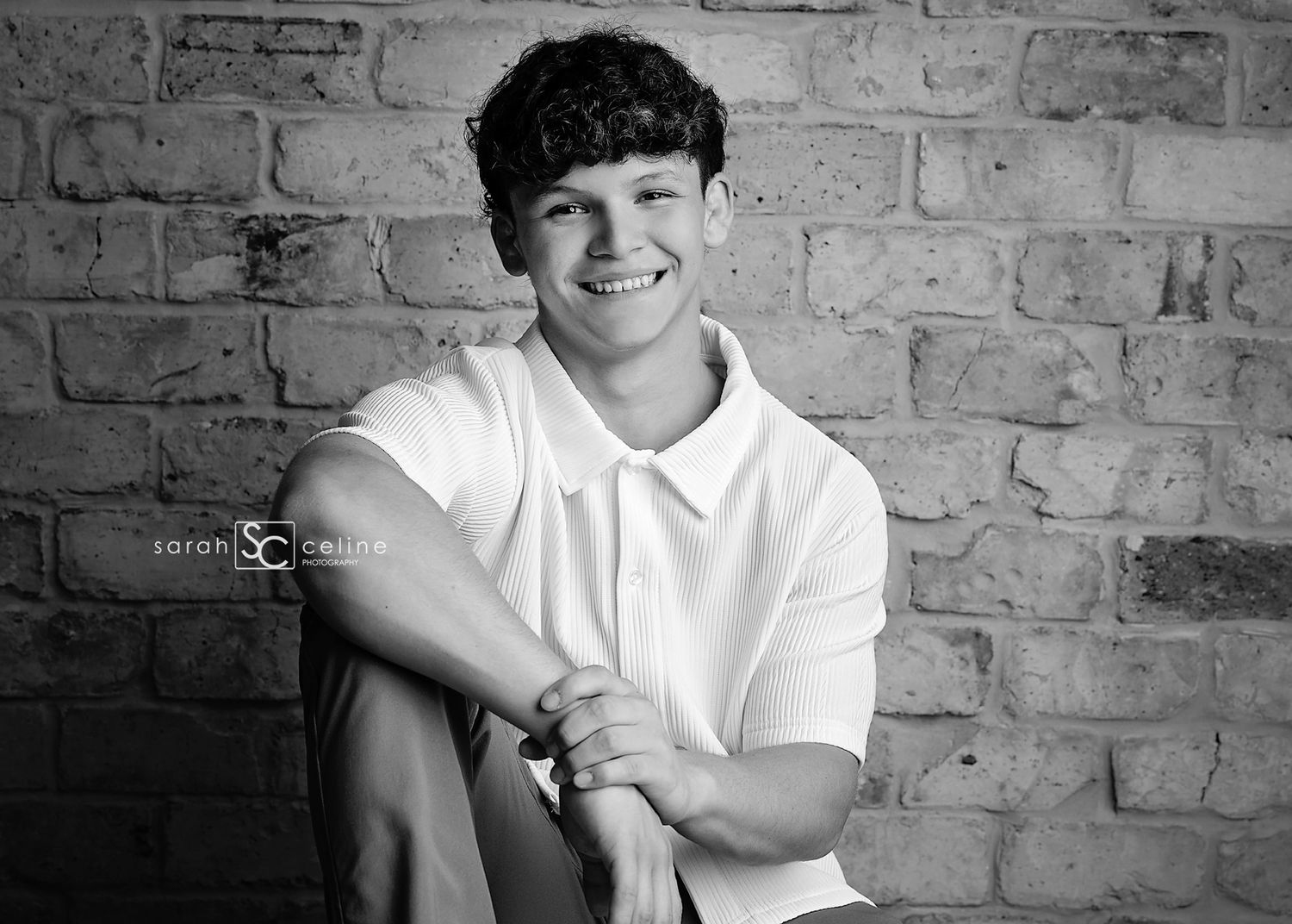 Black and white portrait of a smiling person in a white polo shirt against a brick wall background.