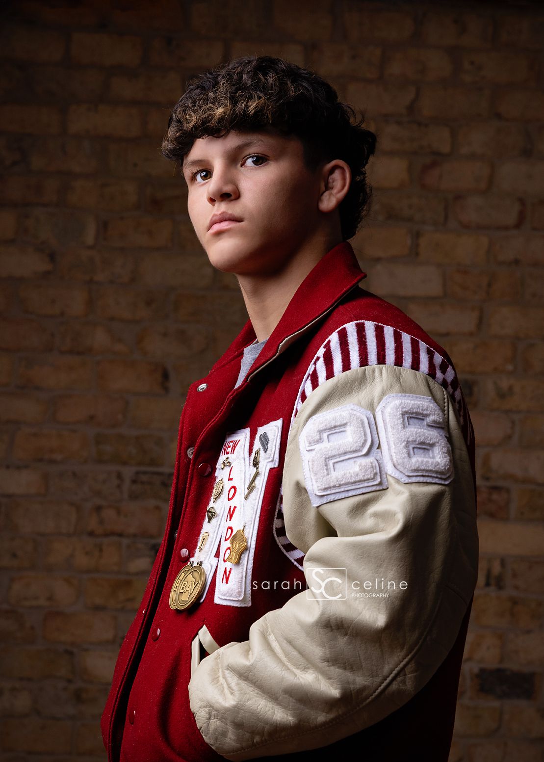 Portrait of a person wearing a red and white varsity letterman jacket against exposed brick.