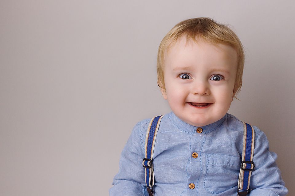 Baby wearing light blue overalls and suspenders holds a white tissue paper with a joyful expression.