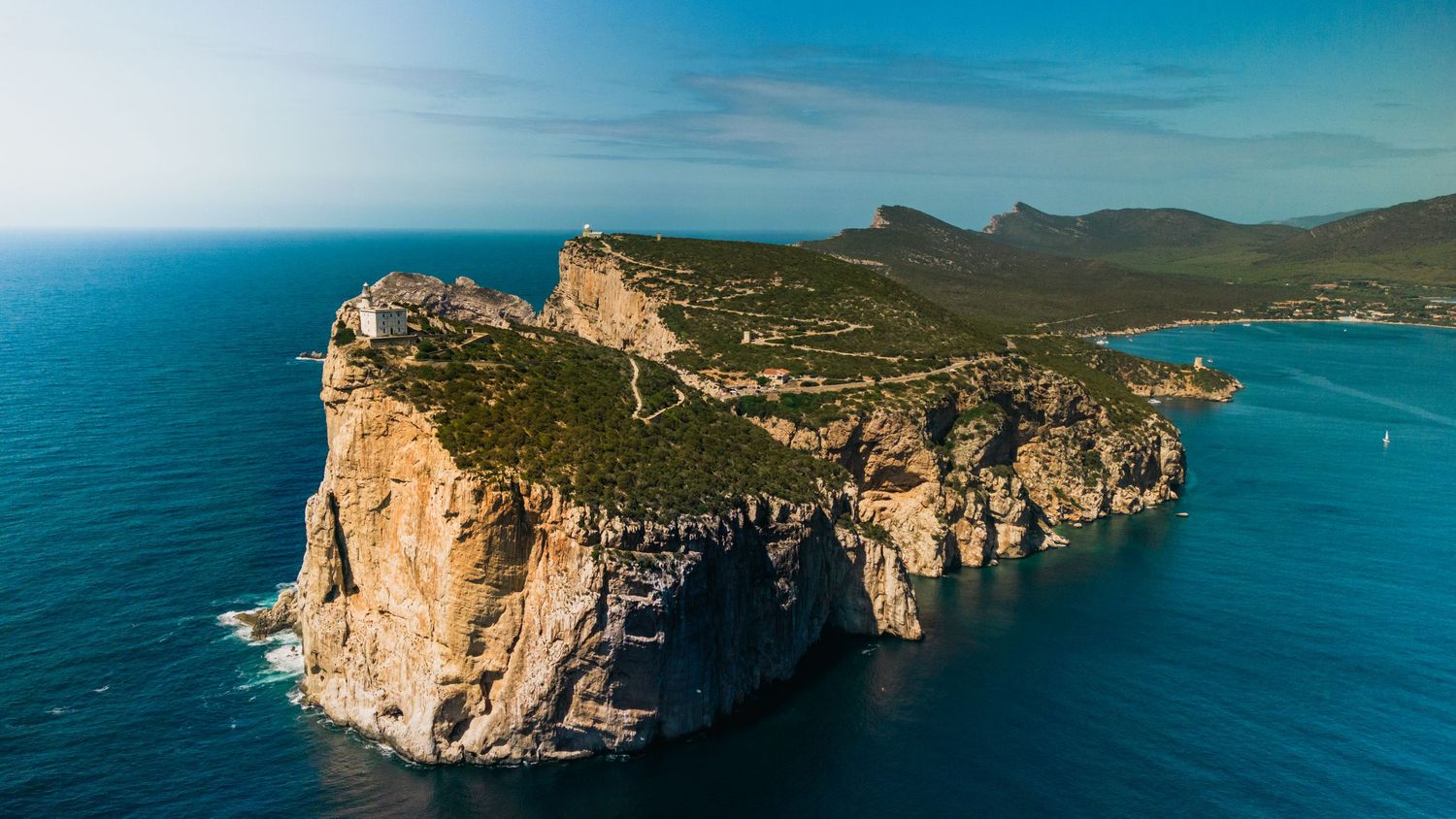 Panoramic coastal landscape with dramatic limestone cliffs rising from turquoise Mediterranean waters under blue sky.