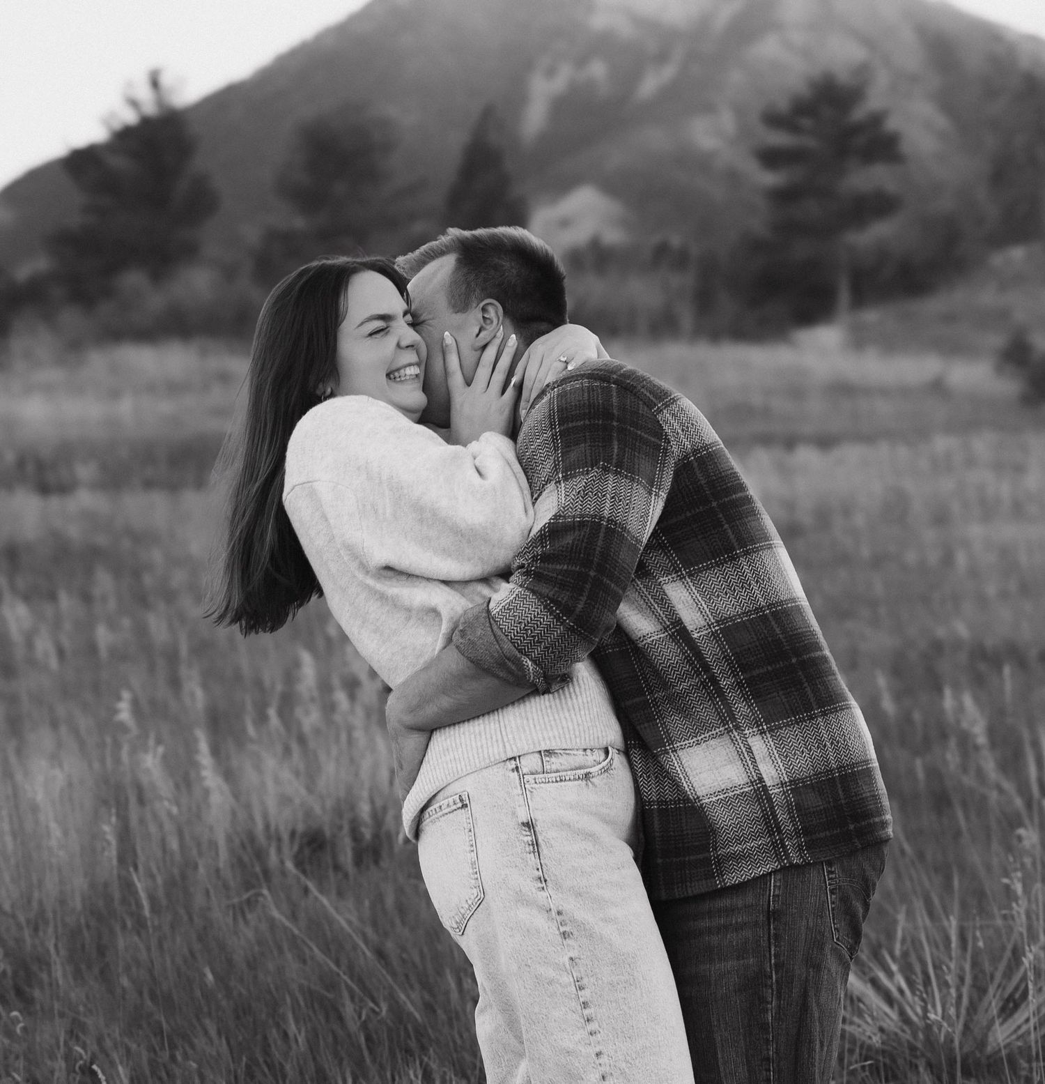 A couple in casual attire embraces and laughs together in a sunlit meadow with tall grass and mountains in the distance.