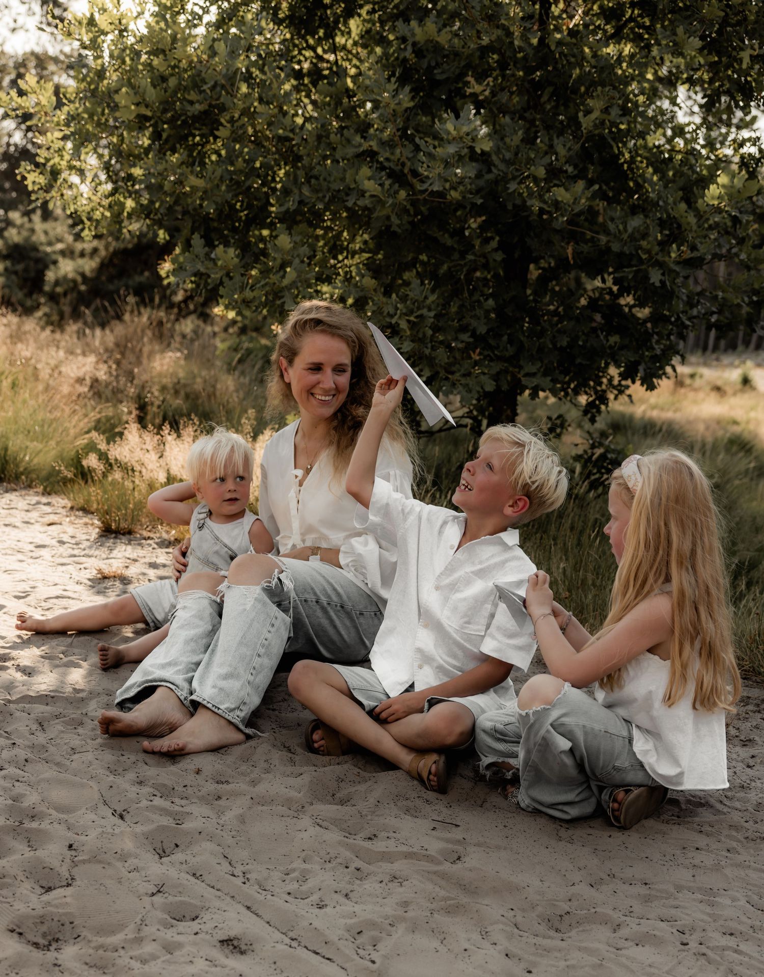 Family relaxing together on sandy ground under trees in warm sunlight.