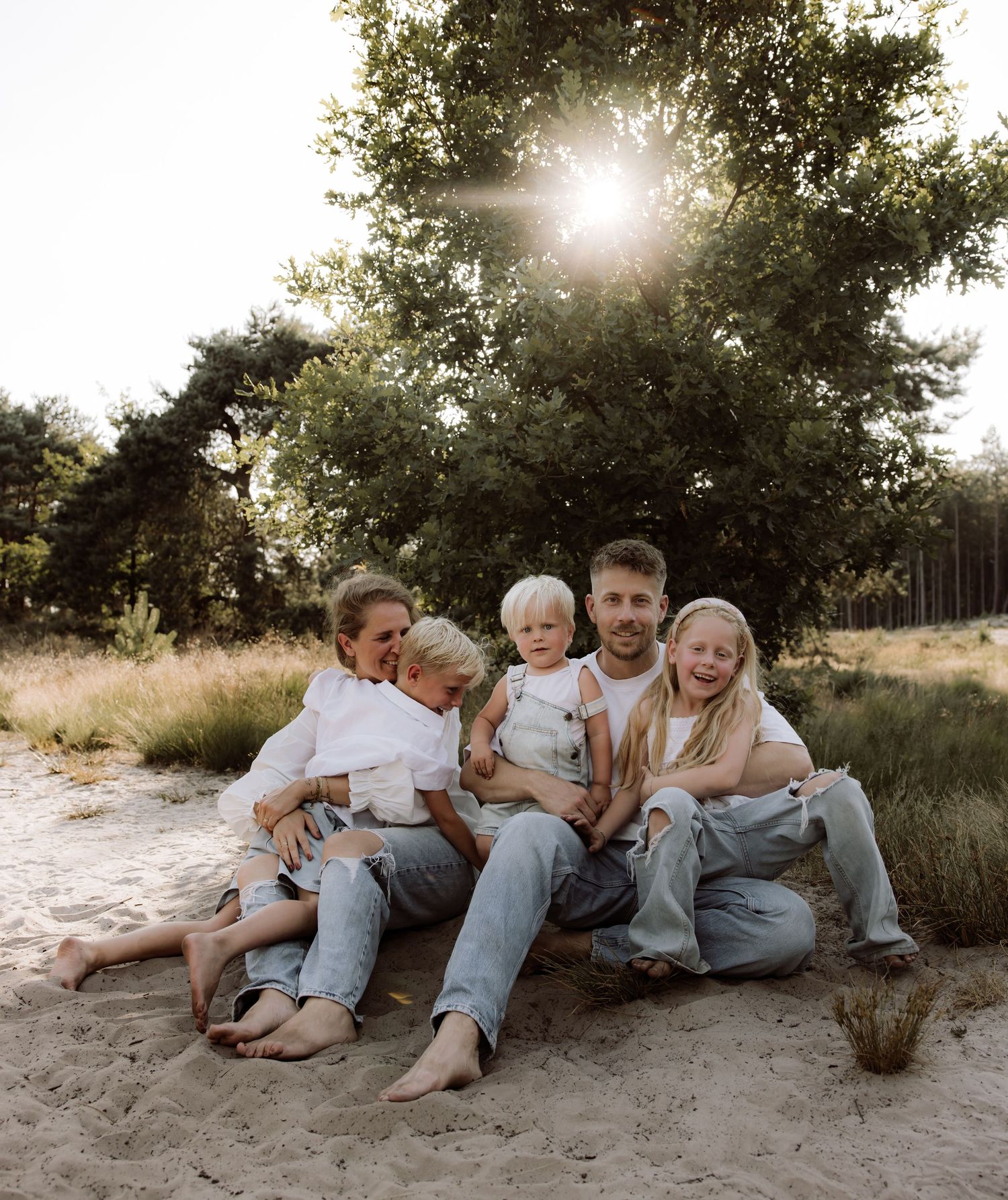 Family lounging together on sandy beach at sunset with trees in background.