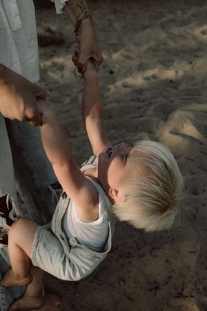 A young child in casual wear being lifted up while playing outdoors on a sandy surface.