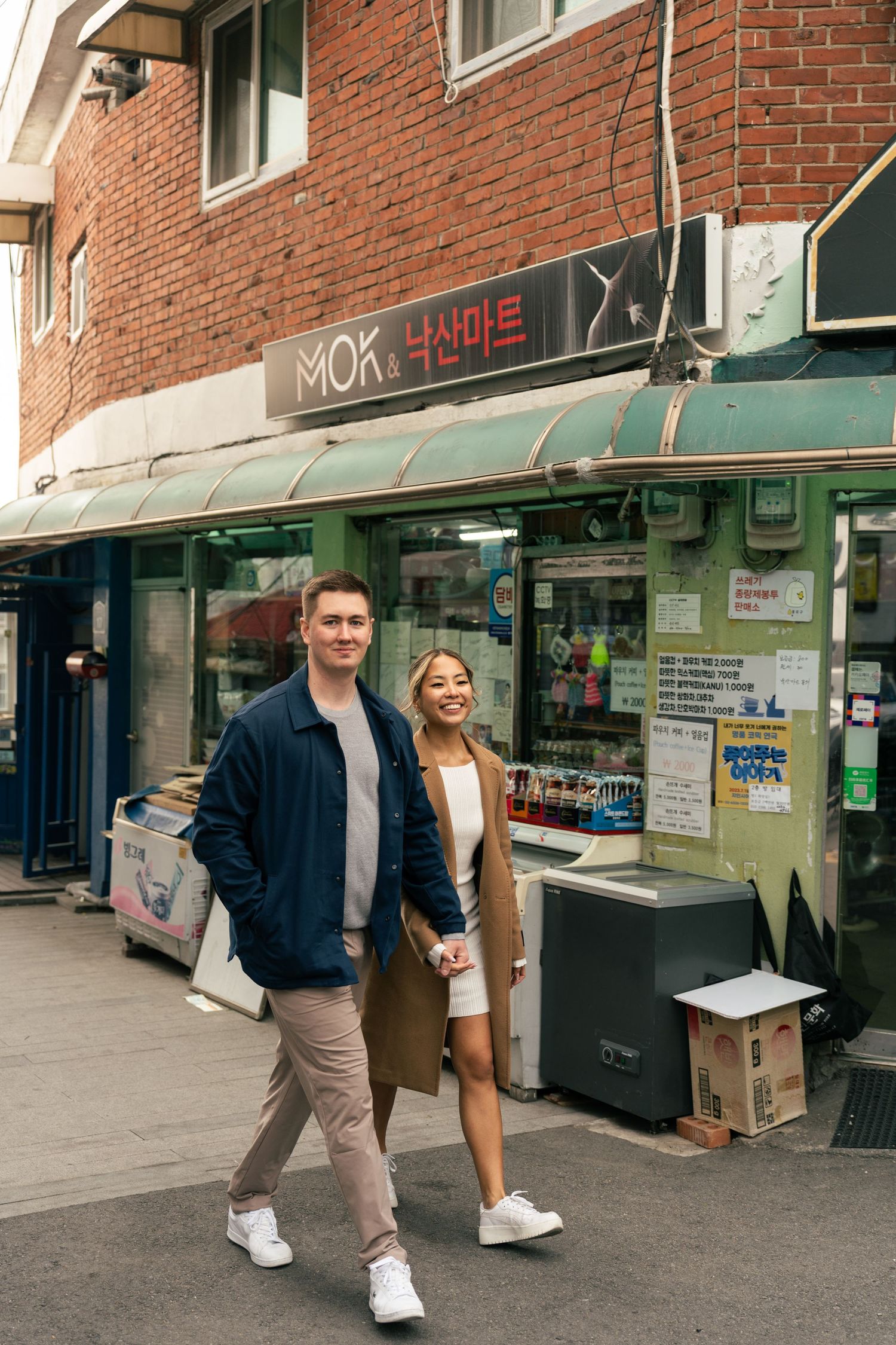 A couple walks past storefronts with brick buildings and retail signs in an urban shopping area.