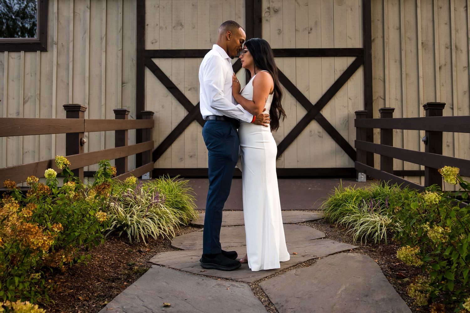 A couple embraces while standing on a wooden deck surrounded by landscaped gardens and rustic architecture.