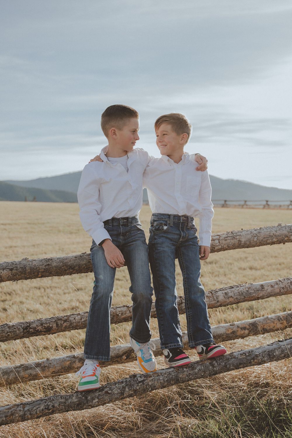 Two children in white shirts sitting on wooden fence rails with mountains in background.