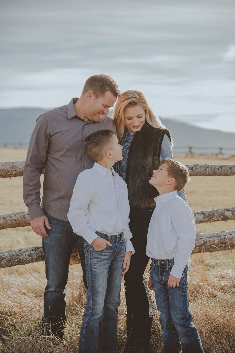 Family gathered close together by wooden fence with mountain landscape behind them.