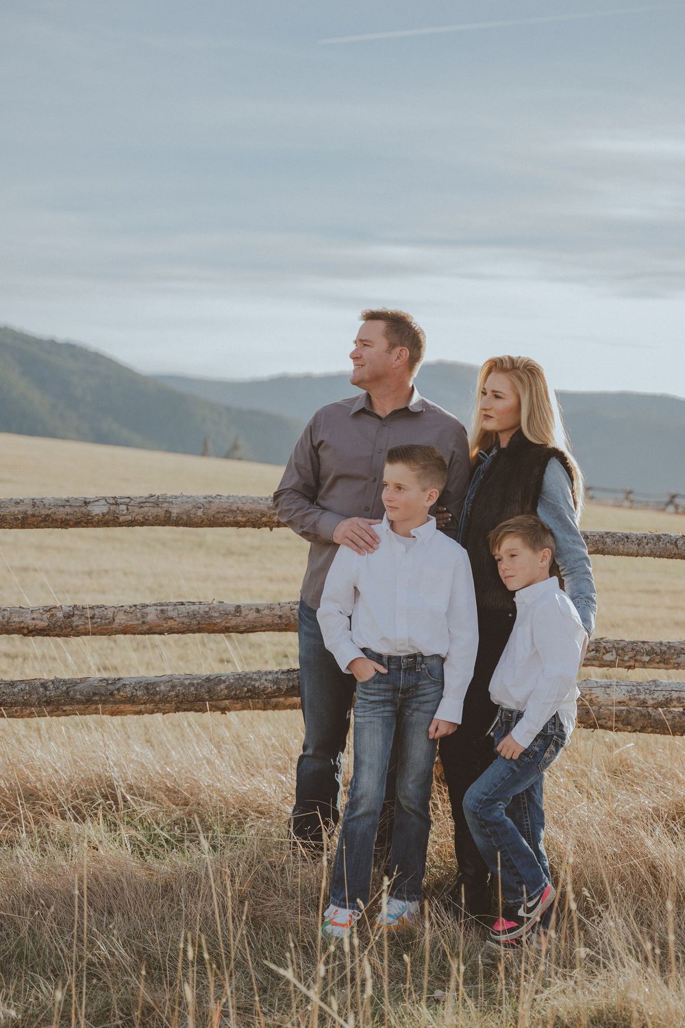 Family portrait in casual attire standing by wooden fence with mountains in background.