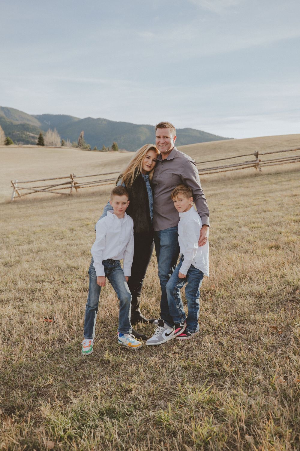 Family playing and laughing together in a grassy field with wooden fence and mountains.