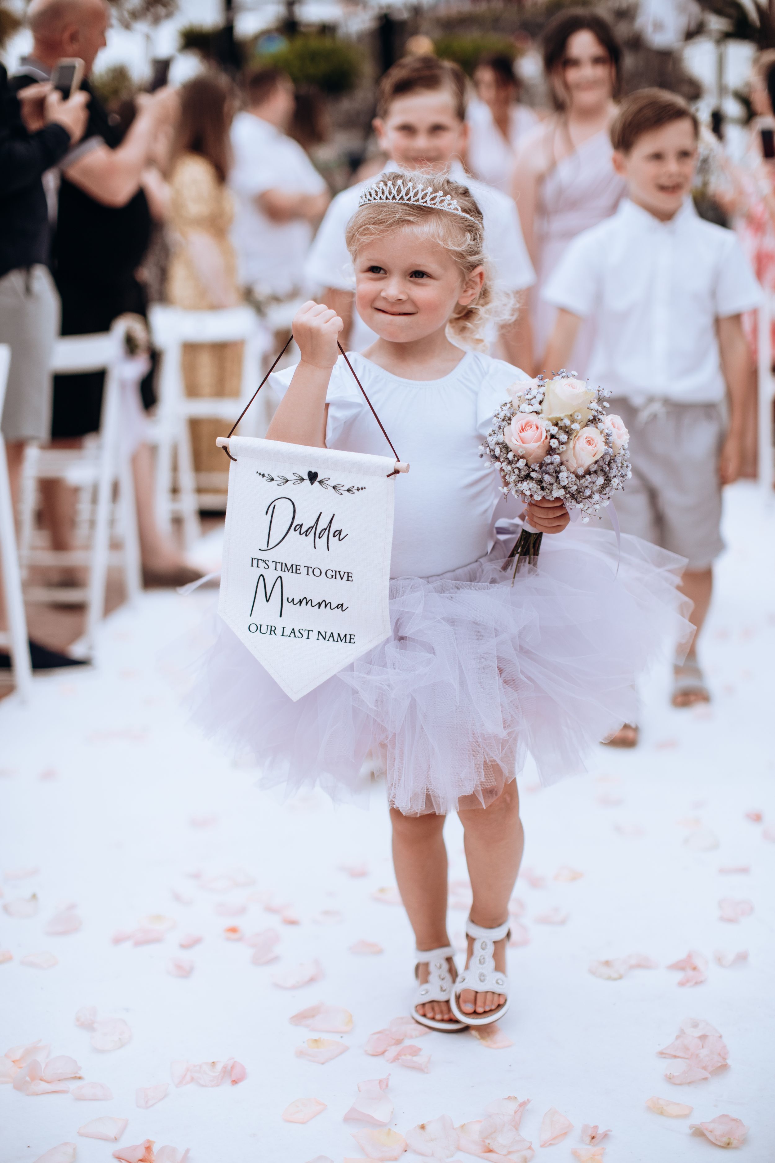 Young flower girls in white dresses with signs walking down aisle at outdoor wedding ceremony.