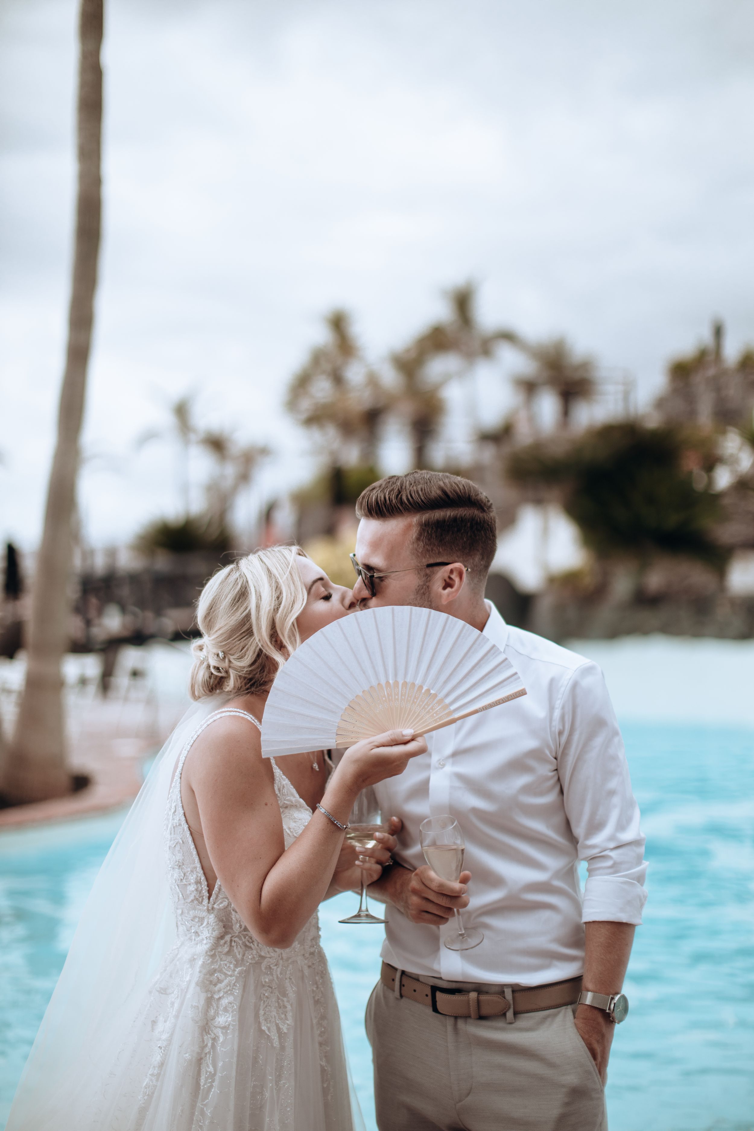 Couple shares romantic moment with white fans at beachside wedding by turquoise pool and palm trees.