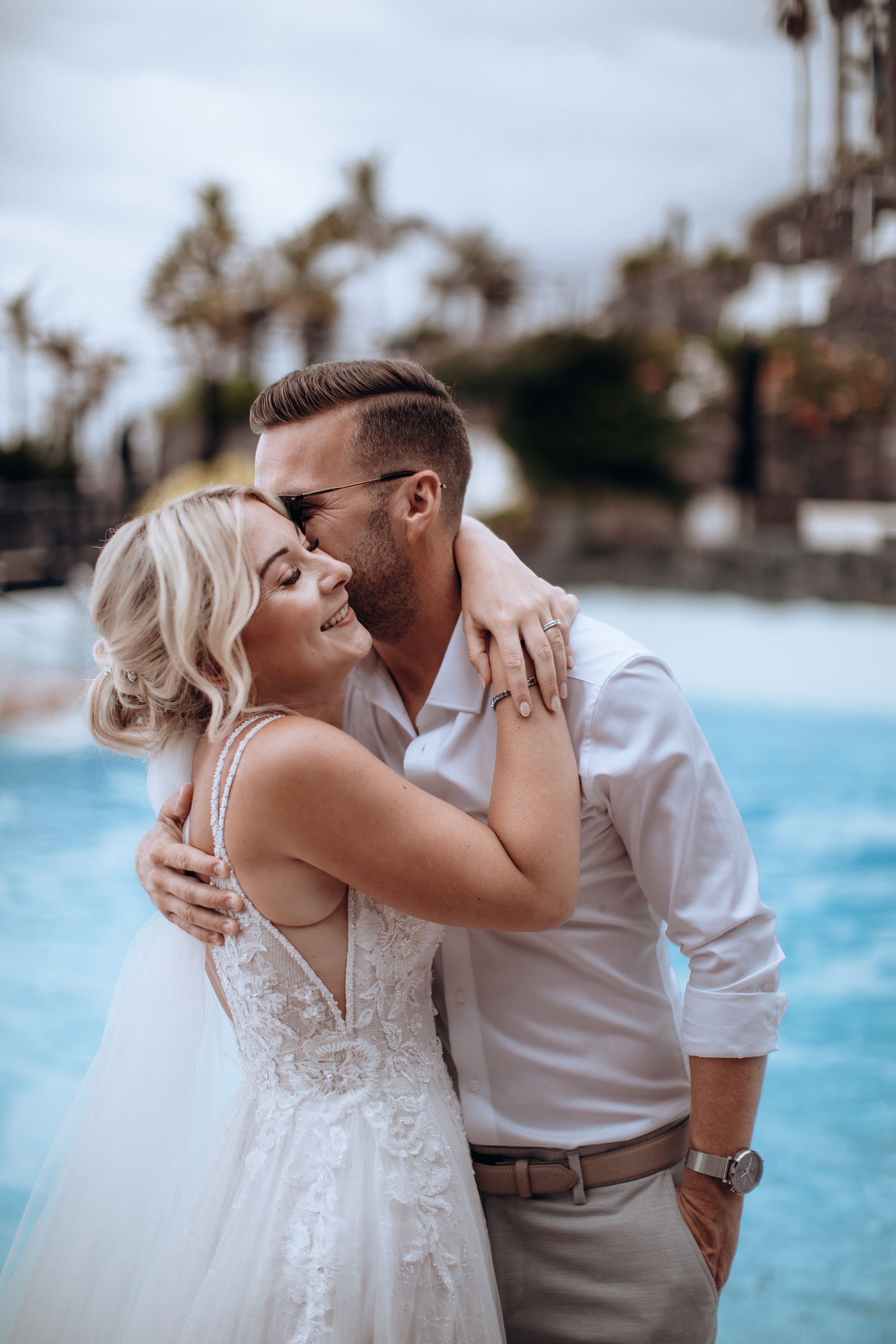 Romantic wedding couple embracing by tropical pool with palm trees in background.