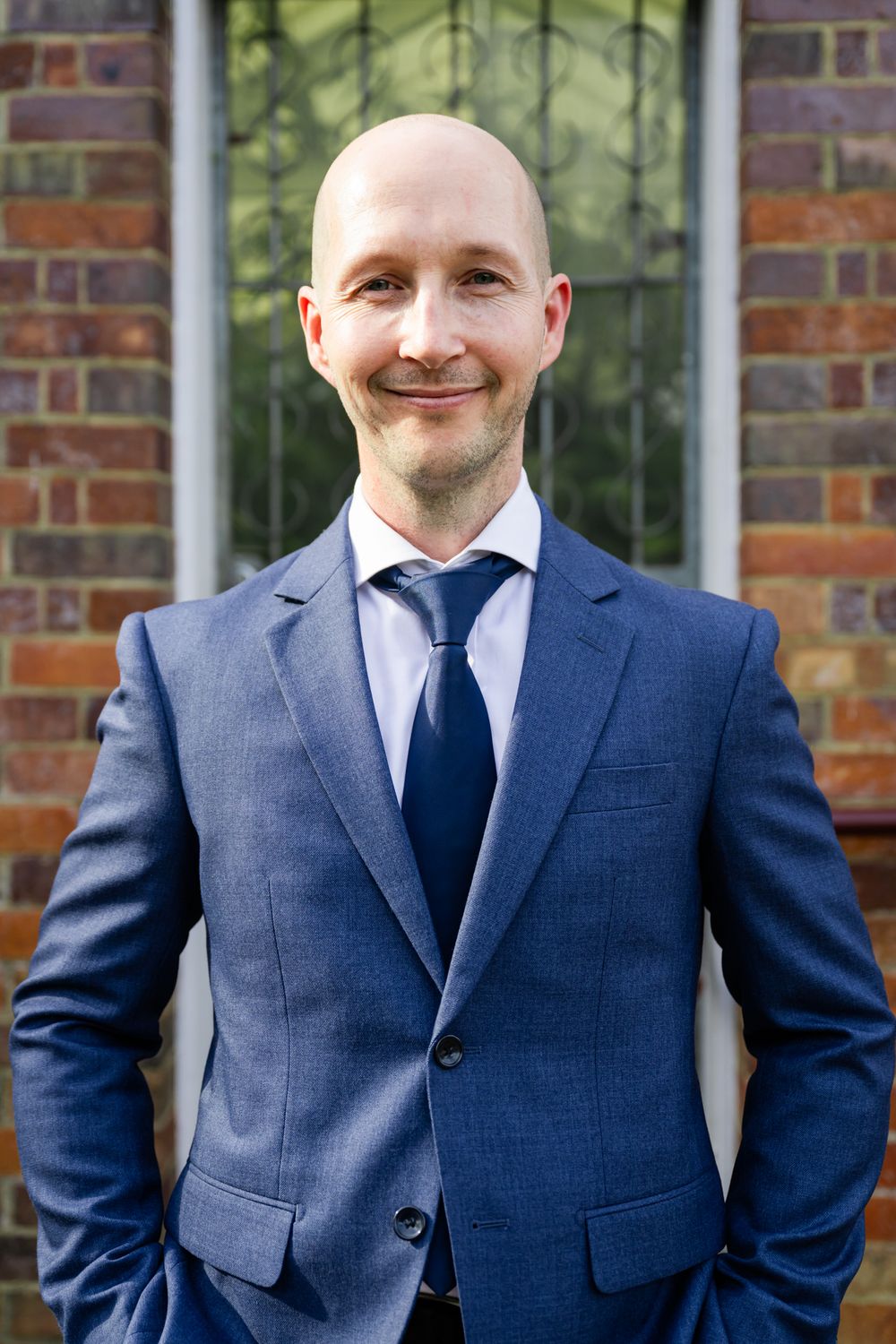Professional portrait of a person wearing a navy blue suit against a brick wall with greenery in the background.