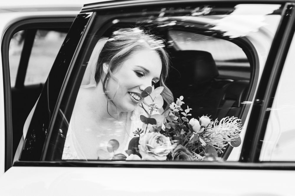 Bride in white dress smiles while sitting in classic car with wedding bouquet in hand.