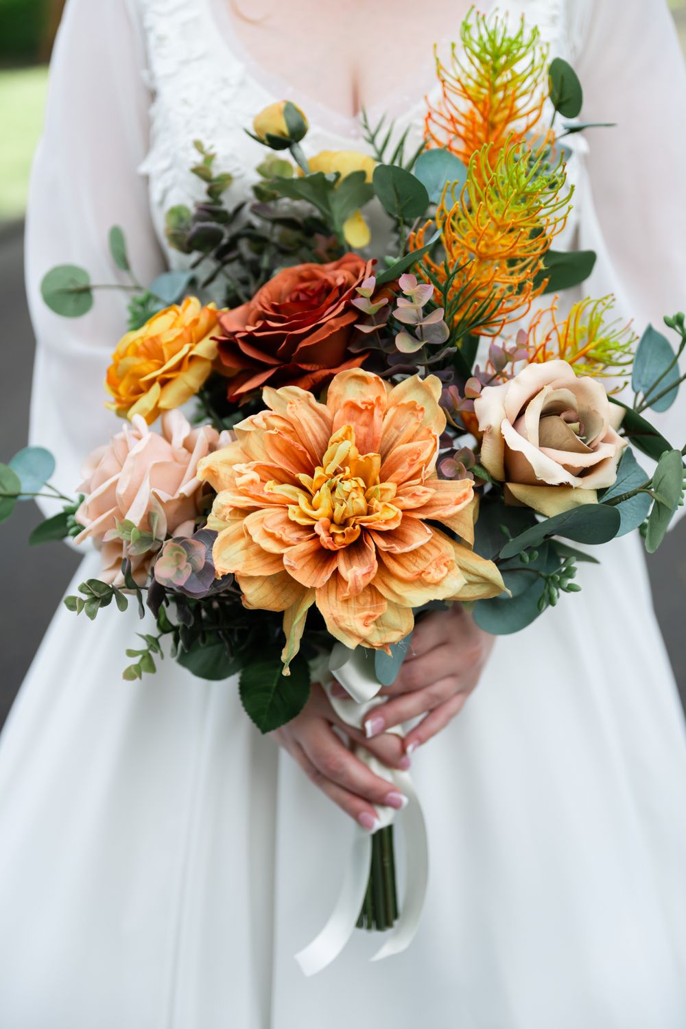 A fall-themed bridal bouquet featuring orange dahlias, roses, and greenery against a white wedding dress.