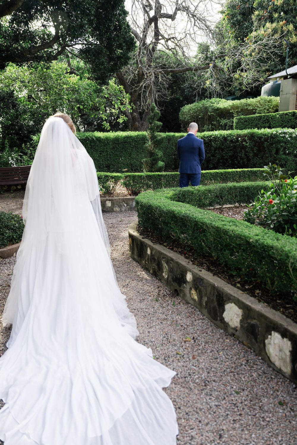A wedding couple stands in a manicured garden with tiered hedges and gravel paths during their first look moment.