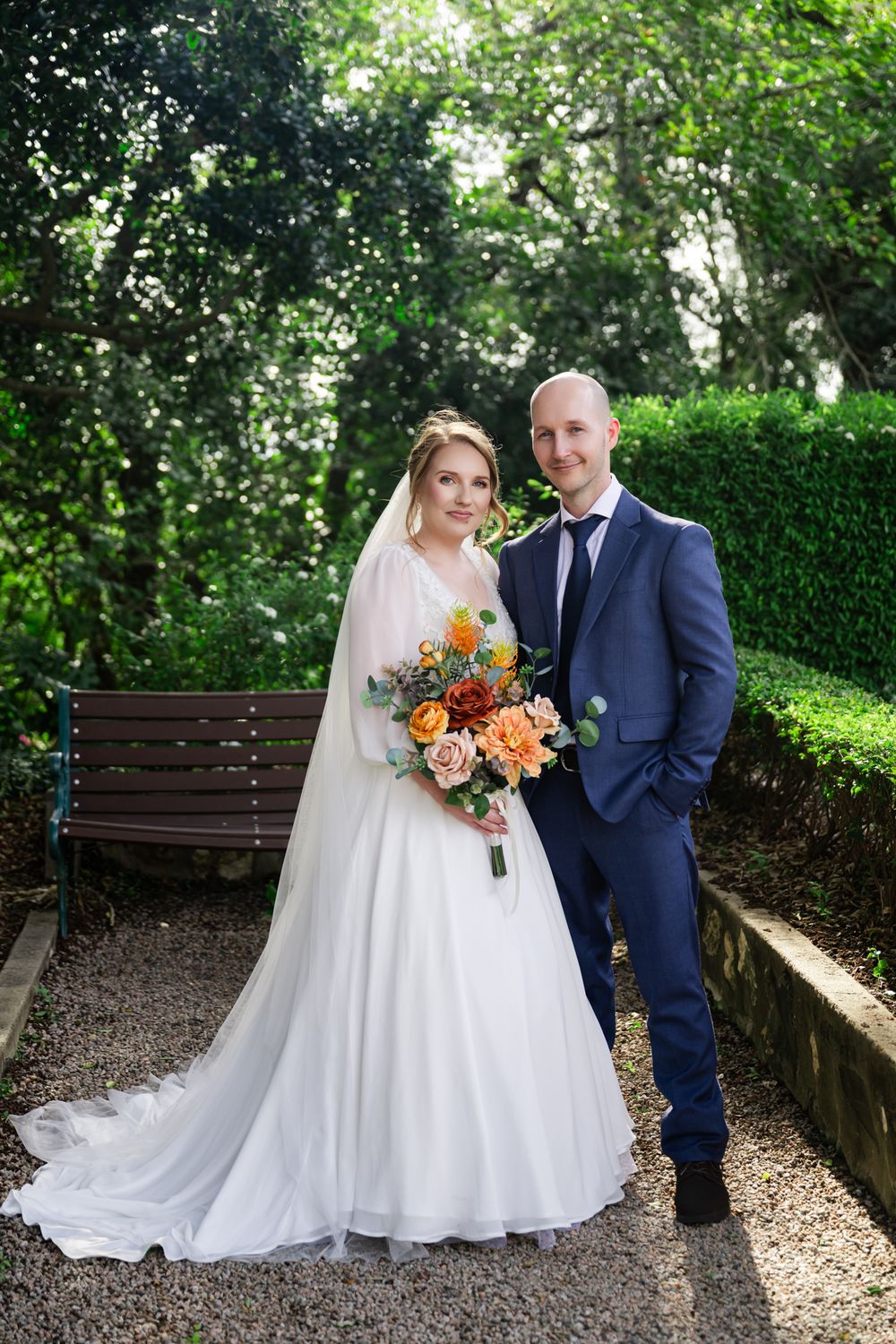 A couple in wedding attire poses together in a lush garden setting with greenery and a wooden bench.