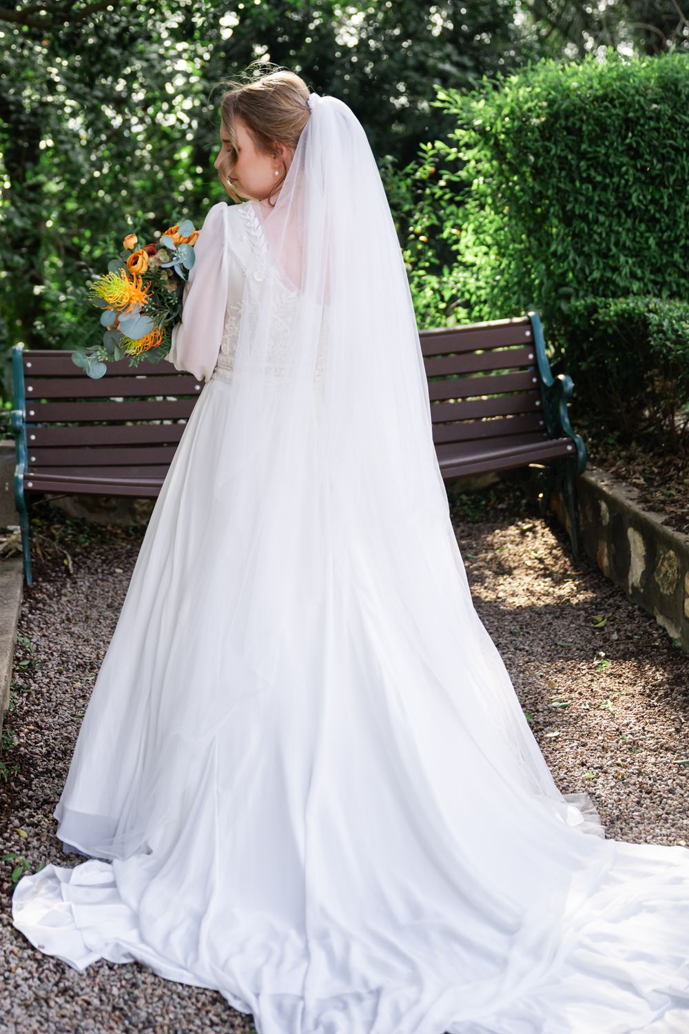 A flowing white wedding dress with a long train is photographed from multiple angles on a garden bench.
