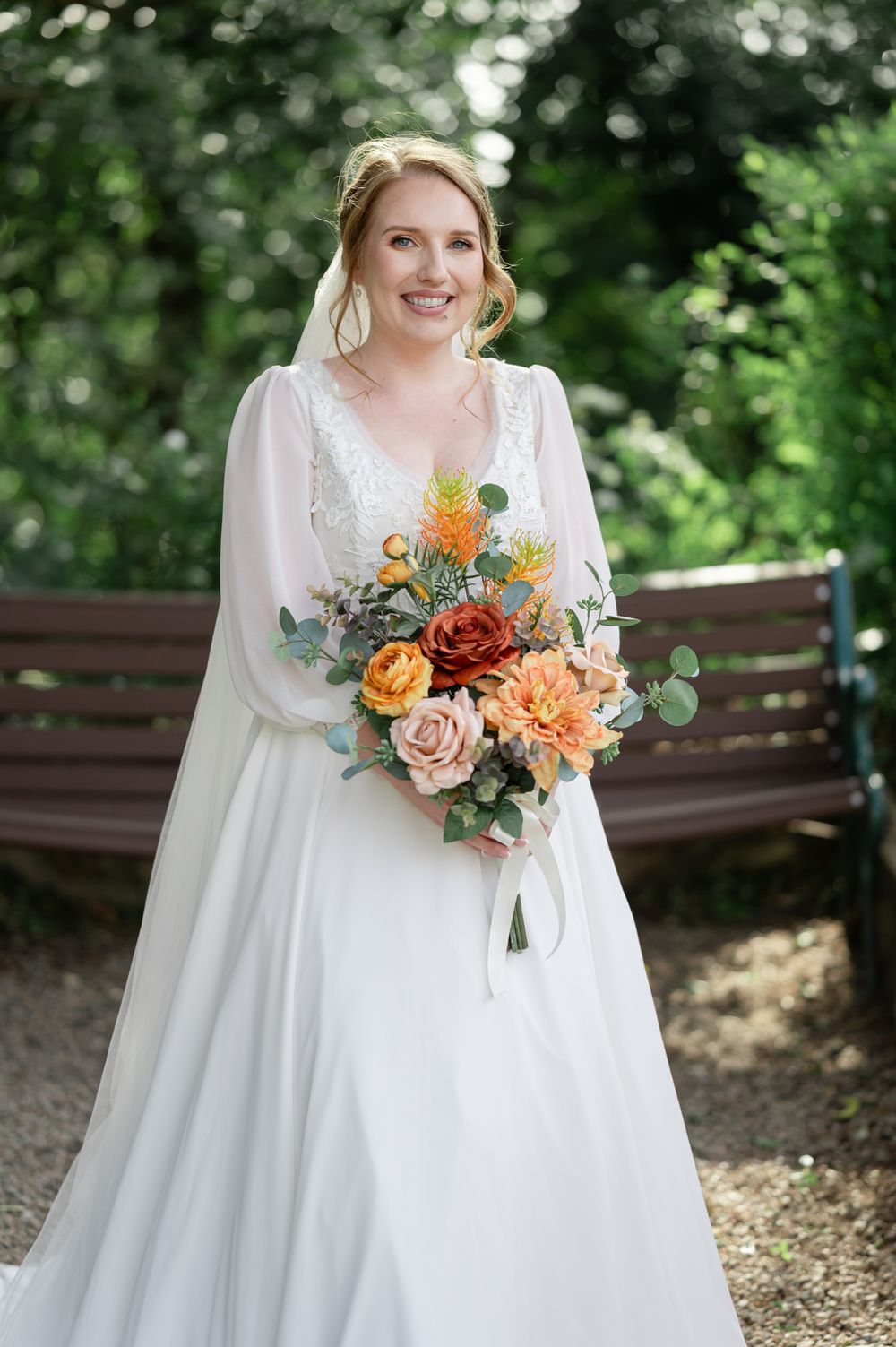 Bride in flowing white dress holds vibrant orange and peach bouquet in outdoor garden setting.