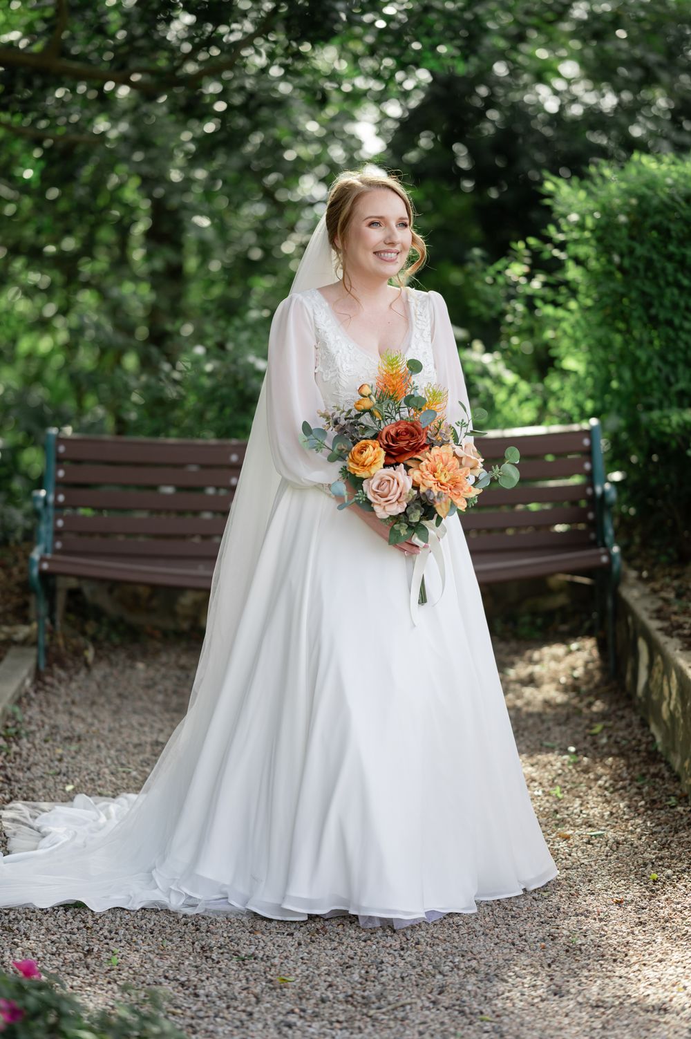 Bride in elegant white gown poses with colorful bouquet on garden path with wooden bench.