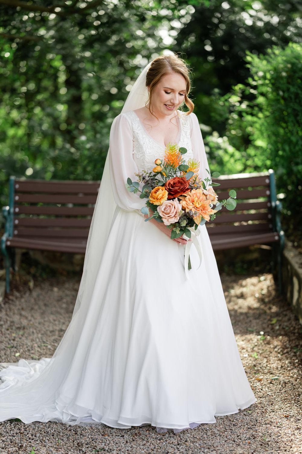 Elegant bride in long-sleeved white gown holds vibrant orange and peach bouquet on gravel path surrounded by greenery.